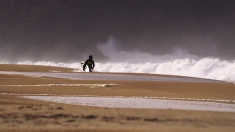 Man-walks-on-an-empty-beach-in-wetsuit-carrying-a-surfboard