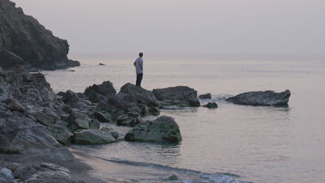 Lone-man-steps-onto-rock-stretching-his-arms-above-his-head-looking-at-sea-as-sun-rises-above-horizon