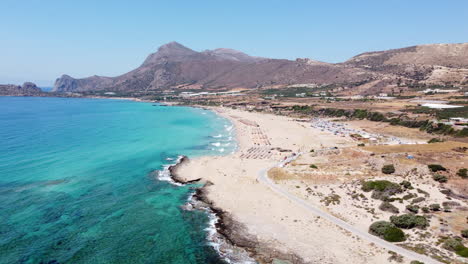 Aerial-view-orbiting-idyllic-Falassarna-beach,-Crete-with-tourists-sunbathing-and-swimming-in-Azure-Aegean-sea