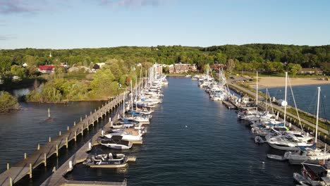 Sunny-day-in-Suttons-Bay-pier-with-many-sailboats-moored,-aerial-drone-view