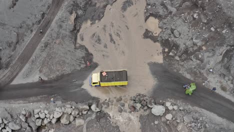 Vehicles-crossing-a-muddy-stream-that-has-flowed-over-a-dirt-road-after-volcanic-aftermath,-aerial-view