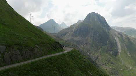 Aerial-Flying-Along-Winding-Mountain-Road-Grimsel-Pass-With-Reveal-Of-Wind-Turbines-On-Ridge-Line-In-Background
