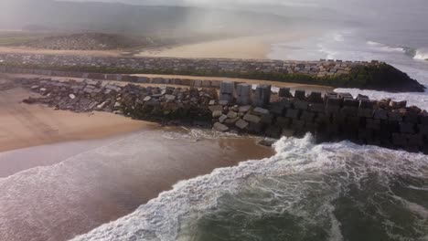 Aerial-view-of-crashing-waves-against-rocky-wall-at-sandy-beach-with-river-mouth-in-Nazaré,-Portugal---Rio-Alcoa,Leira
