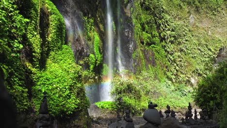 Rock-cairns-stacked-at-the-base-of-a-tropical-waterfall-that-creates-a-rainbow-in-the-canyon-of-a-mystical-rainforest,-East-Java,-Slow-motion-panning-up
