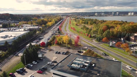 Drone-footage-captures-an-aerial-view-of-an-urban-landscape-bustling-with-traffic-and-activity,-featuring-a-busy-road,-commercial-buildings,-and-autumn-foliage-bathed-in-soft-sunlight