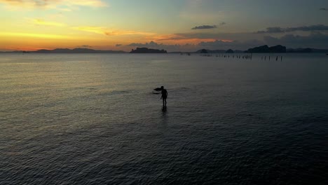 The-silhouette-of-a-young-boy-wandering-and-stumbling-in-the-shallow-sea-waters-of-scenic-coastline-during-sunset,-aerial-pullback-shot