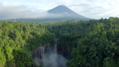 Aerial-view-of-a-volcano-sitting-above-a-large-exotic-waterfall-in-a-tropical-rainforest-at-sunrise,-East-Java-Indonesia