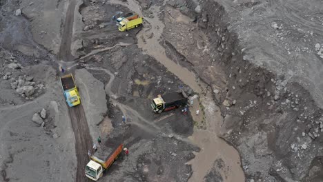 Aerial-view-of-dump-trucks-being-filled-with-black-volcanic-sand-men-pulling-it-from-a-stream,-East-Java-Indonesia
