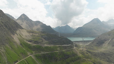 Aerial-View-Of-Winding-Mountain-Road-Grimsel-Pass-With-Wind-Turbines-On-Ridge-Line-In-Background-Beside-Dam