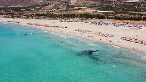 Aerial-view-descending-to-Falassarna-beach-with-tourist-sunbathing-on-sandy-Greek-island-and-swimming-in-transparent-clear-ocean-water