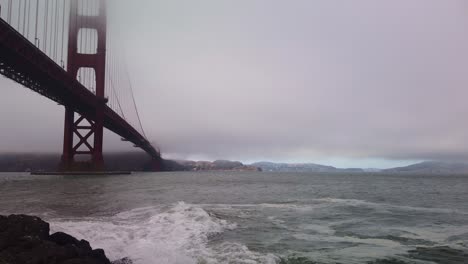 Gimbal-static-shot-of-rough-waves-hitting-the-rocky-shoreline-underneath-the-Golden-Gate-Bridge-on-a-foggy-day-in-San-Francisco,-California