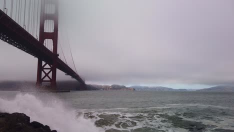 Gimbal-Dröhnt-Nach-Oben,-Aufgenommen-Von-Wellen,-Die-An-Einem-Stürmischen-Tag-In-San-Francisco-Gegen-Die-Felsige-Küste-Am-Fort-Point-Unter-Der-Golden-Gate-Bridge-Krachen
