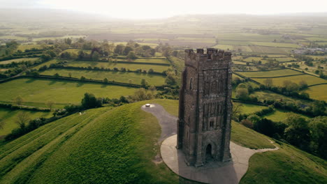 A-drone-shot-circling-Glastonbury-Tor,-a-hill-in-Somerset-UK-the-shot-circles-a-tower-at-the-top-of-the-hill-with-fields-in-the-background