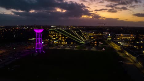 Hyperlapse-of-modern-office-building-with-glowing-water-tower-at-night-time,-aerial