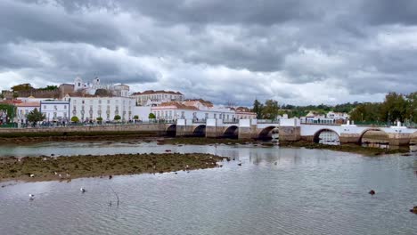 Low-tide-on-the-Galao-River,-people-cross-the-historic-Ponte-Romans-Bridge-with-the-Santa-Maria-Church-on-the-hill-in-the-background-on-a-chilly-spring-day