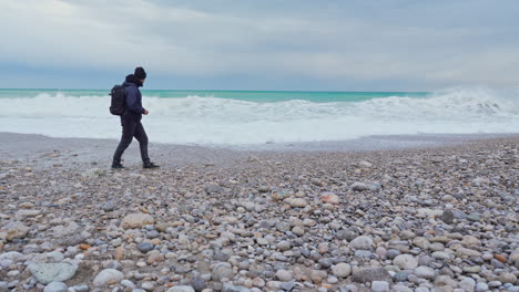 A-man-is-strolling-across-a-stony-beach-while-tumultuous-waves-crash-onto-shore-on-a-gloomy,-overcast-morning