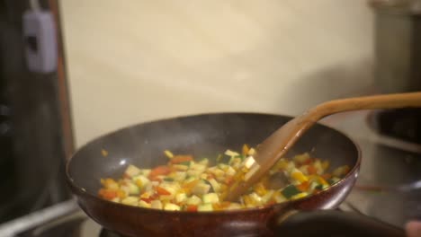 Freshly-chopped-vegetables-gently-fried-in-flat-frying-pan-while-mixed-with-wooden-spatula,-filmed-as-close-up