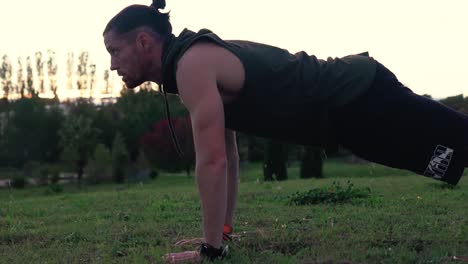 A-muscular-sportsman-is-doing-push-ups-in-a-park,-with-the-sunset-behind