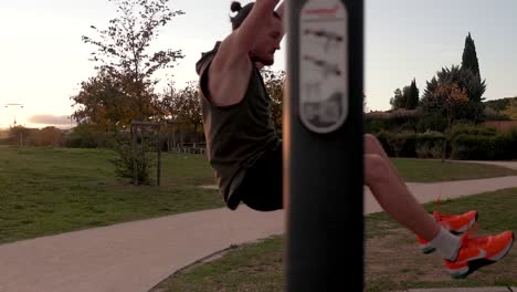 A-muscular-sportsman-is-doing-muscle-ups-in-a-park,-with-the-sunset-behind