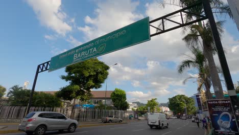 Cars-driving-along-the-main-avenue-of-Las-Mercedes,-as-they-pass-under-the-notice-that-they-are-entering-the-municipality-of-Baruta-in-Caracas,-Venezuela