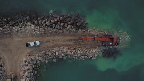 Heavy-machinery-on-breakwater-in-time,-Long-Reach-Hitachi-ZAXIS,-vehicle-and-truck-with-large-stones,-top-down-view