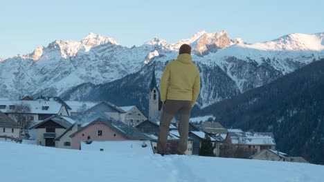 Young-caucasian-man-in-yellow-jacket-and-beanie-stopping-to-see-view-of-small-mountain-village-and-church-in-Guarda,-Switzerland
