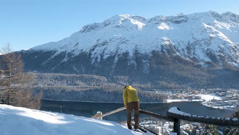 Caucasian-young-man-with-a-yellow-jacket-and-beanie-enjoying-the-view-of-a-lake,-town-and-mountains-on-a-sunny-winter-day-in-St