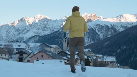 Young-caucasian-man-in-yellow-jacket-and-beanie-running-and-stopping-to-see-view-of-small-mountain-village-and-church-in-Guarda,-Switzerland