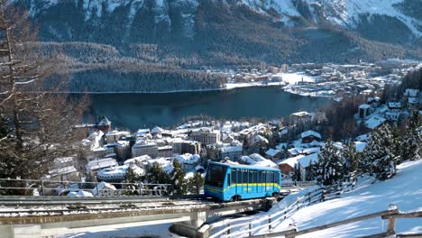 Blue-cable-car-going-up-with-town,-houses,-lake,-trees-and-mountains-in-the-background-in-St