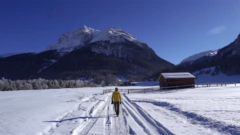 Young-caucasian-man-with-a-yellow-jacket-and-a-beanie-walking-through-snow-covered-winter-landscape-with-a-wooden-cabin