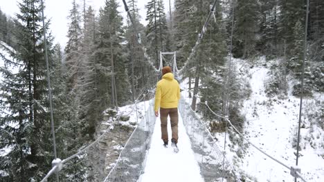 Young-caucasian-man-in-yellow-winter-jacket-walks-across-a-hanging-bridge-in-a-forest-during-winter-while-snowing