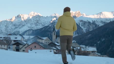Young-caucasian-man-in-yellow-jacket-and-beanie-running-and-stopping-to-see-view-of-small-mountain-village-and-church-in-Guarda,-Switzerland