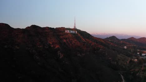 Reverse-pullback-aerial-shot-of-the-Hollywood-Sign-at-sunset-with-mountains-covered-in-snow-in-the-background