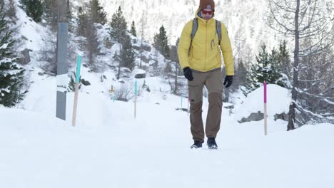 Kaukasischer-Mann-Mit-Rucksack-Und-Gelber-Jacke,-Der-Auf-Einem-Schneebedeckten-Weg-In-Einem-Wald-Auf-Die-Kamera-Zuläuft