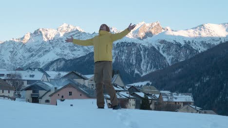 Young-caucasian-man-in-yellow-jacket-and-beanie-stopping-to-see-view-of-small-mountain-village-and-church-and-lifting-his-hands-up-in-Guarda,-Switzerland