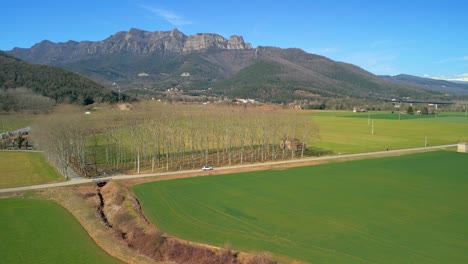 Drone-tracking-a-white-car-on-a-narrow-road-in-an-agricultural-landscape-with-cultivated-green-fields-and-mountains-in-the-background
