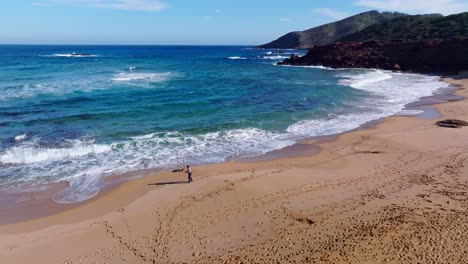 Filmische-Luftaufnahmen-Einer-Person,-Die-Mit-Ihrem-Hund-Am-Strand-In-Cala-Pilar,-Menorca,-Spanien,-Spazieren-Geht