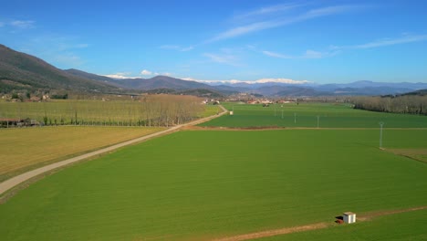 Drone-aerial-footage-of-a-narrow-road-with-cultivated-green-fields-on-the-sides-and-snow-capped-mountains-in-the-background,-forward-flight