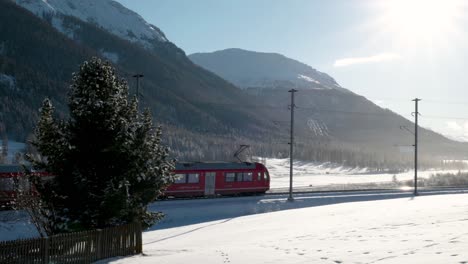 Roter-Zug-Fährt-An-Einem-Sonnigen-Tag-Durch-Schneebedeckte-Landschaft-Mit-Bergen-Dahinter