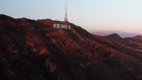 Aerial-close-up-panning-shot-of-the-Hollywood-Sign-at-sunset-with-snow--covered-mountains-in-the-background