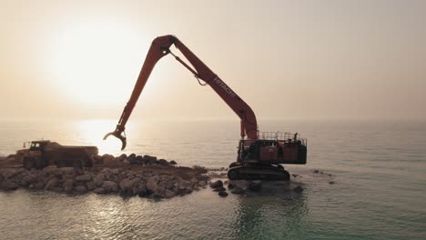 Excavator-machine-takes-stones-from-a-truck-to-build-a-stone-pier-in-the-sea,-sunset-parallax-shot