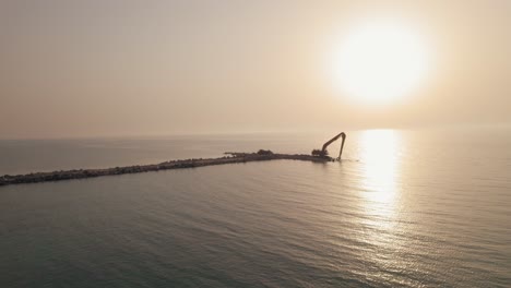 Long-Reach-Excavator-machine-waiting-to-receive-stones-to-build-a-breakwater-in-the-sea,-magical-shot-during-sunset