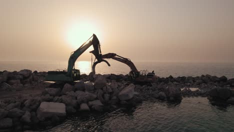 Sunset-silhouette-of-two-excavators-standing-on-a-breakwater-they-constructed