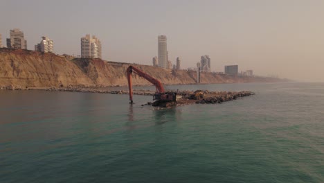 Single-excavator-machine-builds-a-breakwater-at-sea,-pull-back-shot-at-sunset