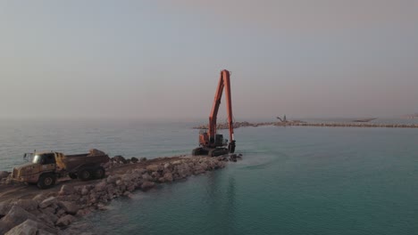 Long-Reach-Excavator-machine-receives-stones-to-build-a-stone-pier-in-the-sea,-sunset-parallax-shot