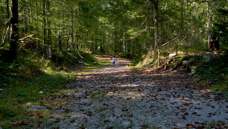 Girl-running-in-forest-behind-mother,-playing-in-woods,-foothpath-with-autumn-foliage,-shot-from-behind,-facing-away,-4k
