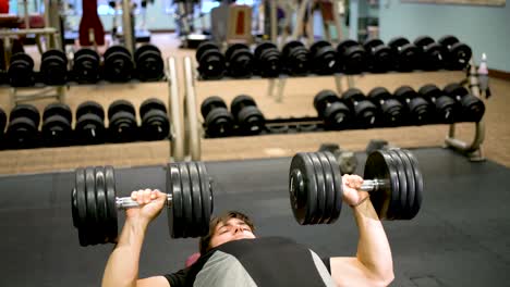 Front-shot-of-teen-bodybuilder-lies-down-on-bench-and-presses-large,-heavy-dumbbells-in-a-twisting-motion
