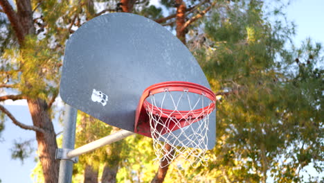 An-old-basketball-hoop-with-a-metal-backboard,-orange-rim-and-net-in-an-empty-park-court-at-sunrise