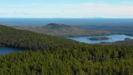 Aerial-drone-shot-flying-over-a-forested-ridge-to-reveal-blue-mountain-lakes-in-the-forest-along-a-mountain-range-as-summer-ends-and-the-season-changes-to-fall-in-Maine