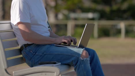 Closeup-Man-Typing-on-Laptop-sitting-on-metal-park-bench-outdoors